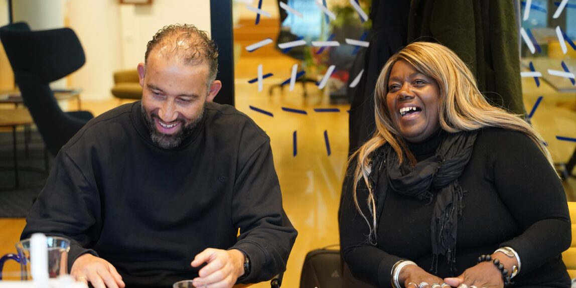Een Afro-Surinaamse vrouw met lang blond haar en een Arabische man met een volle baard lachen hartelijk aan een workshoptafel.