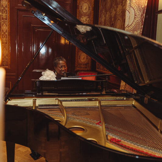 Liesbeth Peroti zingt en begeleidt haarzelf op een pianovleugel. De ruimte is sfeervol verlicht door een kandelaar vol brandende kaarsen.